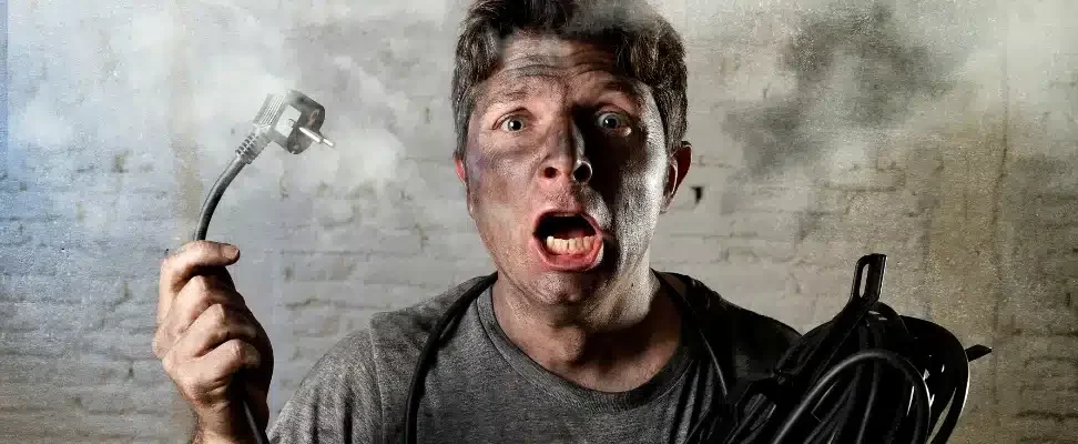 Visuel d'un homme tenant un câble en main, il a le visage couvert de suie et il y a de la fumée en arrière plan. Ce visuel illustre de manière caricaturale et humoristique un accident électrique.